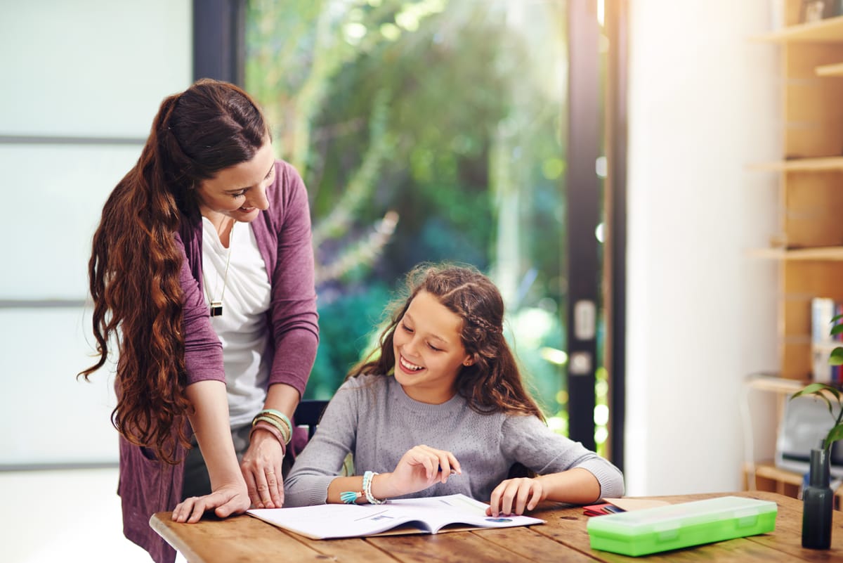 Una madre inclinada sobre la mesa ayudando a su hija sonriente con las tareas escolares en casa